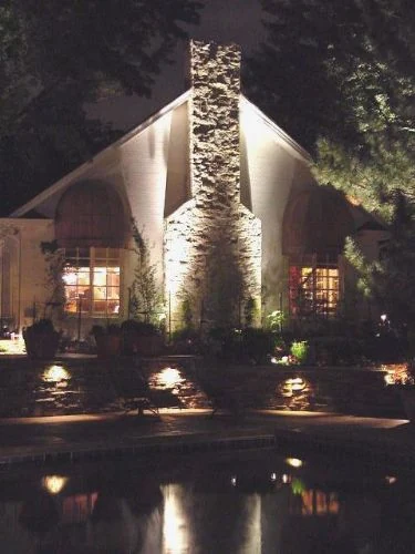 A tall stone chimney and gable lit at night and reflected in the water below.