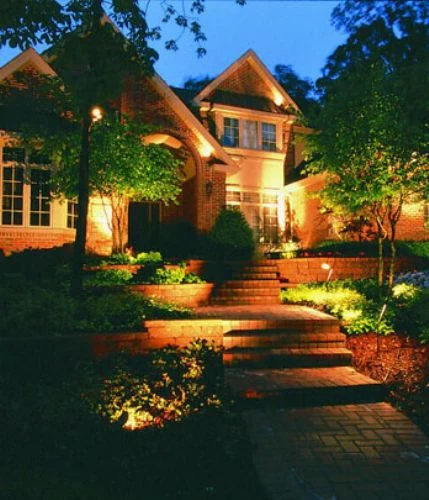 A lit walk and steps leading up to a brick house with lit trees behind it.